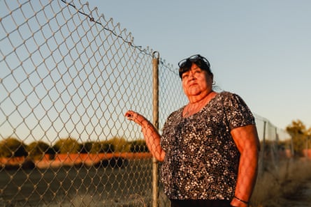 A larger Latina woman wearing a short-sleeved shirt and with short black hair stands alongside a chain-link fence, which she holds onto with her right hand, looking into the setting sun.