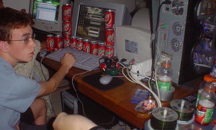 A boy sitting in front of a computer surrounded by cans of drink, and with other computers and paraphernalia on the desk next to him