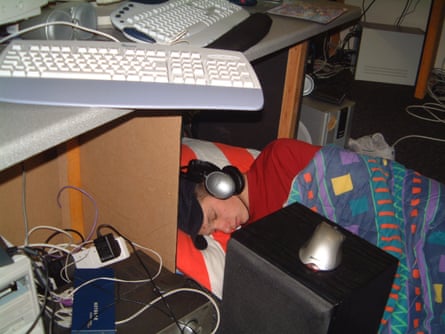 A boy with headphones on asleep in a sleeping bag under a desk with computers on it and in front of it