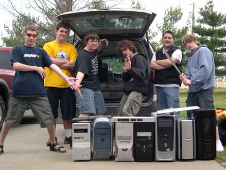 Six teenage boys standing behind the open boot of a large car and pointing at computer hard drives and keyboards on the ground in front of them