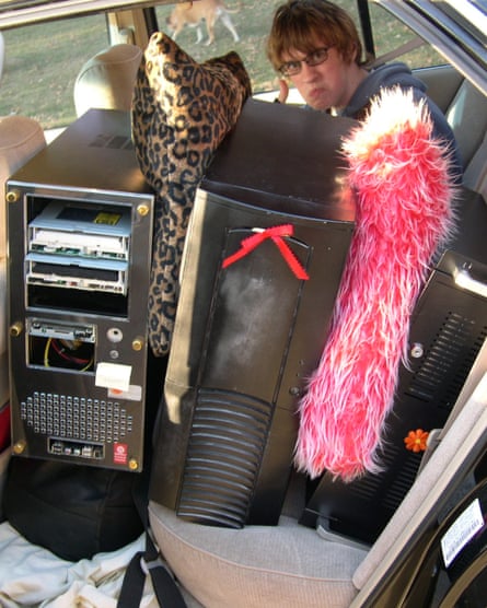 A boy sitting in the back of a car with computer hard drives beside him padded out with a pink fluffy cushion and a leopard print cushion