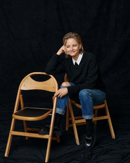 Portrait of a smiling Jodie Foster sat on a chair while resting her foot on another one. Sweater: Cos. Blouse: Theory. Jeans: Foster’s own. Shoes: Gucci. Jewellery: Anita Ko.