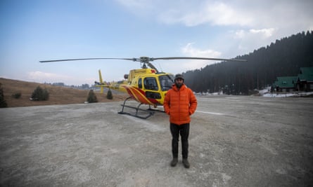 A solemn-looking man in a down jacket stands in front of a yellow helicopter