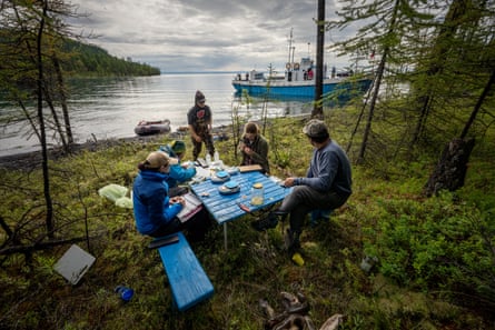 A group of researchers sit around a picnic table on the shore of lake with a boat moored in the background