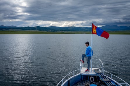 A man stands next to a flag on a pole at the front of a boat on a lake