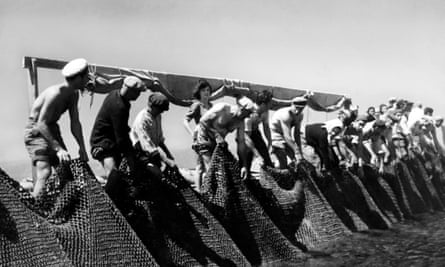 A black and white photo of a line of men holding the edge of a big net, with a few women standing behind them