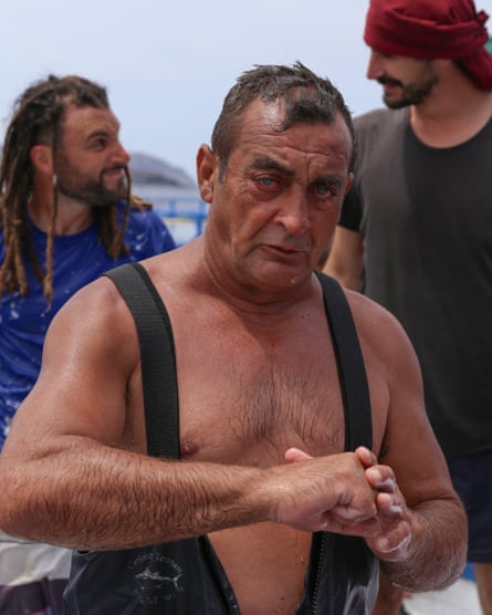 A sun-burnt older man drenched with seawater and with red eyes looks at the camera as two young men behind him chat