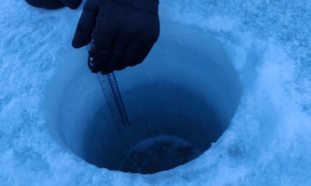 A hand in a mitten holding a ruler in a hole cut through the ice 