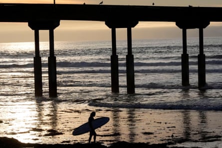 Bright yellow evening sunlight reflects on ocean waves beneath a sky that appears to have clouds on the horizon, with a person holding a surfboard in silhouette alongside the two-story-high pilings of a bridge, also in silhouette.