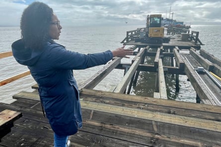 A Black woman with curly shoulder-length hair and wearing a 3/4-length rain jacket and jeans holds her right arm out toward a section of the cement pier she stands on that is only iron cross bars.