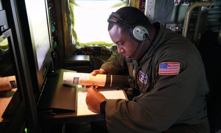 A Black man with short black hair, wearing an olive-colored jacket with an American flag patch on the shoulder, hunches over a desk wearing headphones and holding a small-ish, conical thing, writing with his left hand.