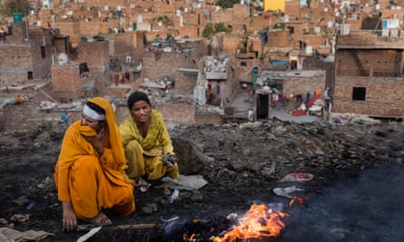 Ragpickers burn wires to extract copper at the Bhalswa landfill site, Delhi, 9 October 2023: two young women, one in an orange sari and headscarf, the other in yellow, squat by flames on the piles of rubbish. The woman in orange is covering her mouth against the smoke