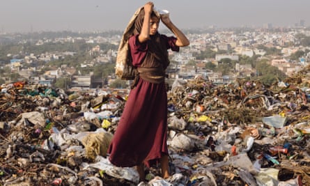 A young girl in a dark red dress and brown headscarf holds a bag filled with rubbish over her head. She is standing on piles of trash with the city seen in the background
