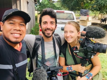 A smiling young Indigenous Brazilian man stands next to a young American man and a young American woman, both holding camera equipment 
