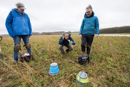 Two researchers stand in a field and one crouches listening to the soil.