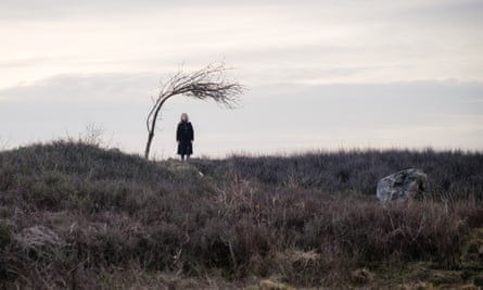 A woman stands on empty moorland with a bare-branched tree bent over her