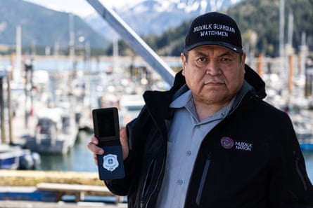 A man wearing a hat and a jacket bearing the words Nuxalk Guardian Watchmen stands holding open a leather wallet with a silver shield badge inside it.