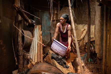 A grim-faced African woman picks up a plastic basket from a tin shack covered in mud 