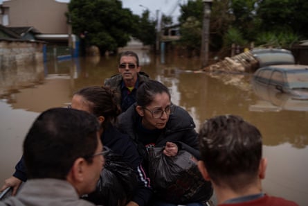 People huddled in a crowded boat, in brown flood waters. A car is submerged just behind them. 