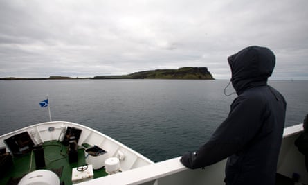 The prow of a small, white ferry boat is seen in the foreground of a view of an expanse of grey sea; a cliff and the edge of an island is seen in the background under a grey, cloudy sky. A person in a black hooded anorak is silhouetted at the front of the boat and seen from behind; their hood is up over their head. 