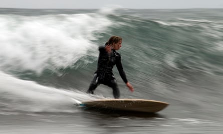 A white woman in a wetsuit leans into a wave a bit higher than her as she surfs 