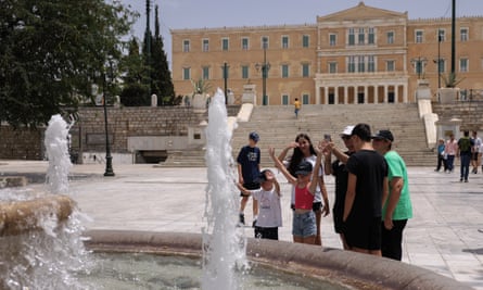 A family group with young children stands next to a fountain; a large classical-style building with pillars at its entrance is seen in the background at the top of a flight of steps across a large stone public square. A boy and girl, both wearing T-shirts and baseball caps, are holding their hands out towards the water.