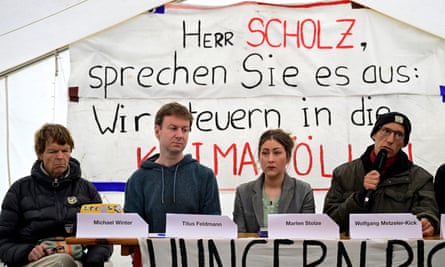 Four people sit on chairs behind a table in a tent at a climate camp. Metzeler-Kick is holding a microphone. A banner behind them, in German, says ‘Mr Scholz, spell it out – we are steering into climate hell’