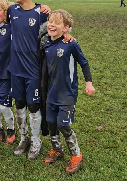Toby in full dark blue football kit celebrates beside teammates in front of a muddy football pitch.