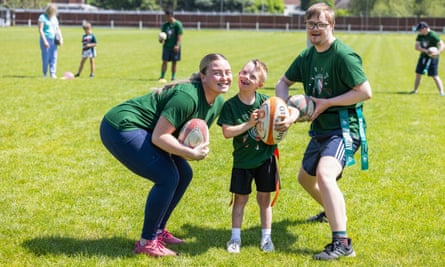 Megan Kirby, the coach, leans down with her face alongside that of Noah, eight, as 22-year-old Harvey stands by; all three wear the green team T-shirt, are holding rugby balls, and are smiling widely