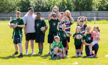 The players pose with professional Leicester Tigers player Nic Dolly, with their coach, Megan Kirby, on the right: the group of boys and girls spans a variety of ages and heights, and they look happy; some are holding rugby balls in the air above their heads