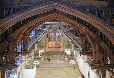Overhead view of the arched medieval beams of Westminster Hall, with hanging scaffolding gantries during refurbishment of the roof in 2019, with stained glass windows in the distance and a few people standing on the stone steps