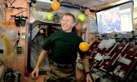 Tim Peake on board the International Space Station with controls and a large screen to his right and pieces of fresh fruit circling around him