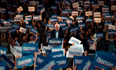 Bernie Sanders stands at a podium in the middle of a rally with people all around him holding ‘Bernie’ posters