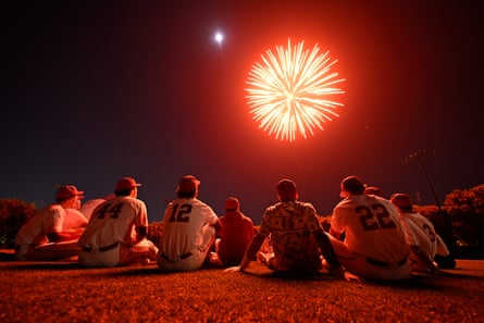 A night sky is lit by the explosion of a red firework, while groups of people sitting on flat ground are illuminated.